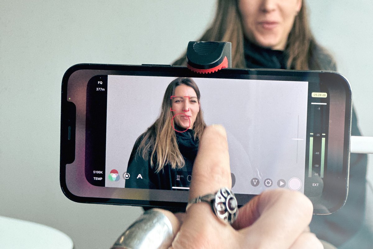 Blick über eine Hand mit Smartphone auf das Display, darauf die befragte Person mit Gesichtserkennung und Tonpegel