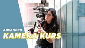 Eine Person mit langen Haaren bedient eine professionelle Videokamera in der Nähe einer Metalltür; der Text lautet "Kamerakurs Advanced November 2023".