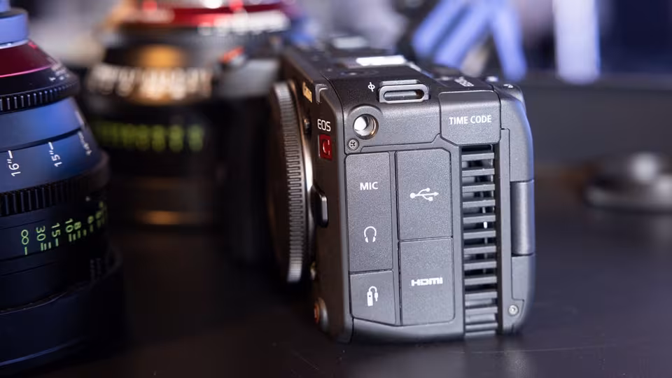 Nahaufnahme der seitlichen Anschlüsse einer Kamera mit den Bezeichnungen MIC, Kopfhörer, USB, HDMI, TIME CODE; im Hintergrund zwei unscharfe Objektive.