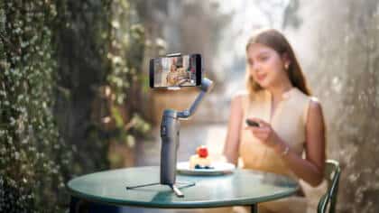 Frau an einem Tisch im Freien mit Dessert, die eine Fernbedienung benutzt, während sie sich selbst mit einem auf einem Stativ montierten Smartphone aufnimmt, dahinter Grünzeug.