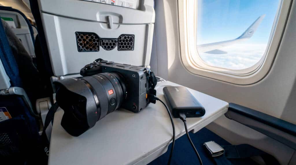Kamera, Festplatte und Powerbank auf einem Flugzeugtisch am Fenster, während draußen Wolken und ein Flügel zu sehen sind.
