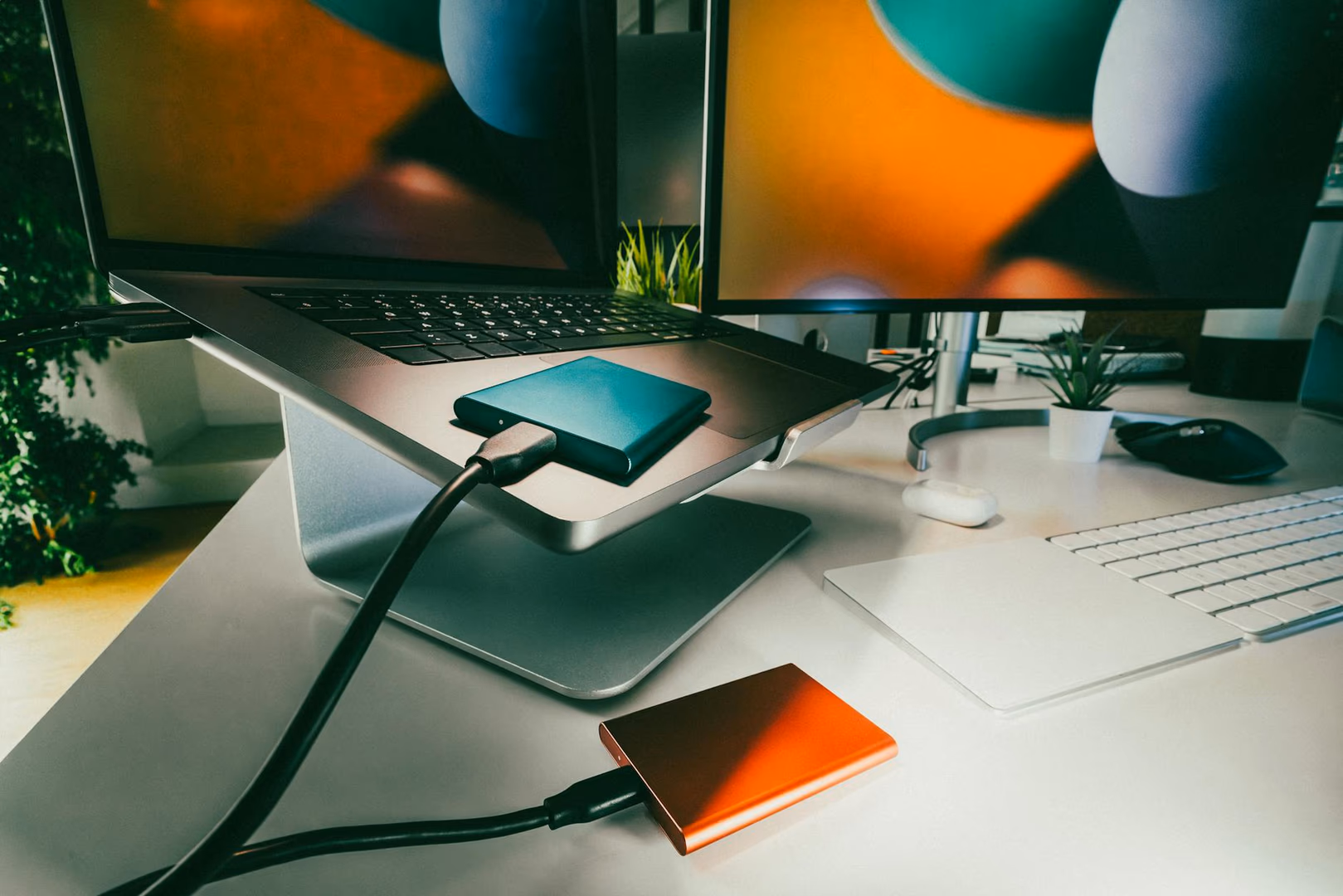 Schreibtisch mit zwei Monitoren, Laptop, externer orange SSD mit Kabel, schwarzer externer Festplatte, Tastatur und Pflanzen im Hintergrund.