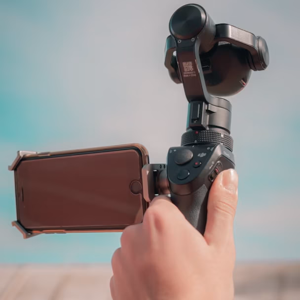 Hand hält eine schwarze kompakte Kamera mit Gimbal-Arm nach oben, vor blauem Himmel und unscharfem Hintergrund.
