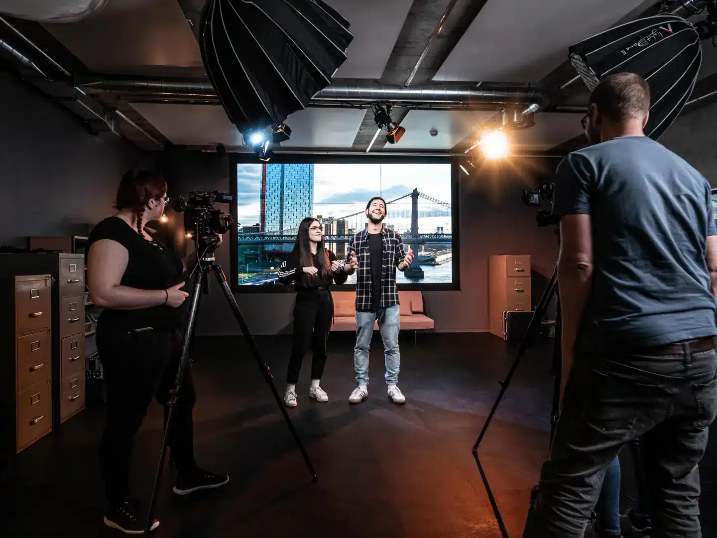 Filmset in einem Studio: Zwei Personen stehen vor einem großen Monitor, der eine Stadtkulisse mit Brücke zeigt. Links und rechts je ein Kameramann an Filmkameras. Im Hintergrund Scheinwerfer, Aktenordner und ein Sofa.