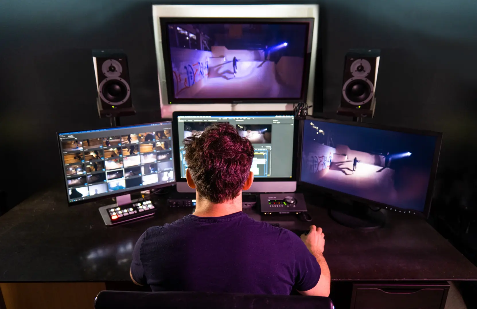 Videoeditor von hinten an einer Schnitt-Suite mit drei Monitoren, Timeline und Media-Bin sichtbar, kalibrierte Studio-Monitore links und rechts