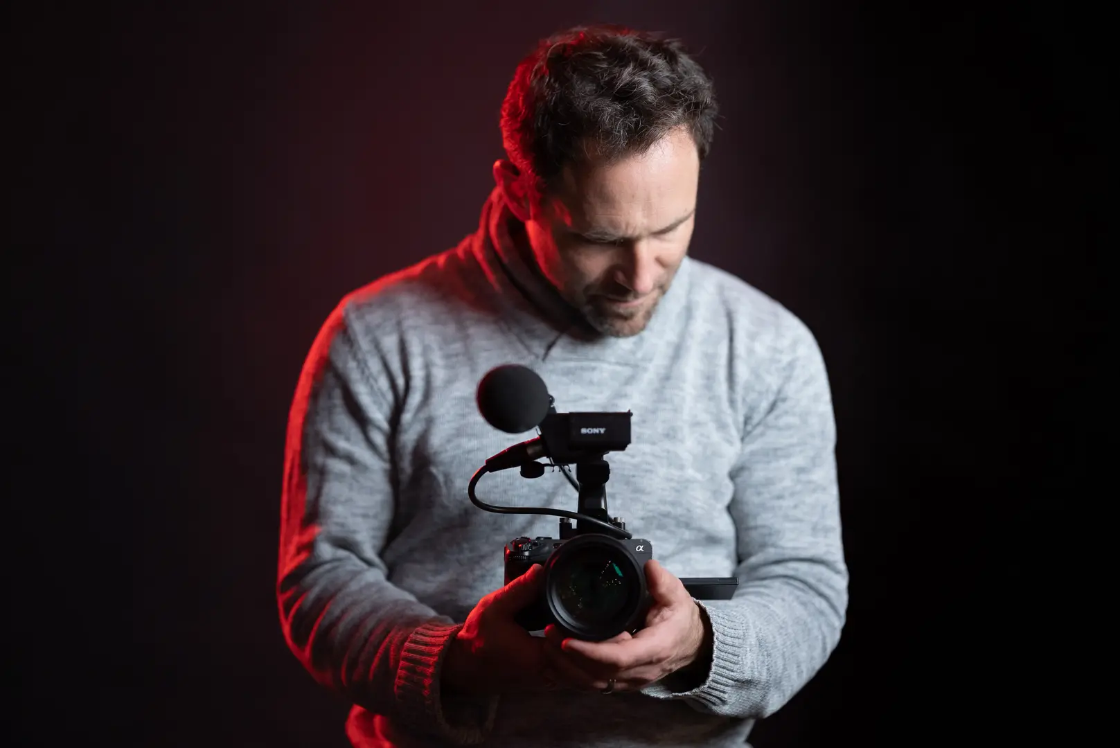 Mann in grauem Pullover hält eine Sony-Videokamera mit aufgesetztem Richtmikrofon – typisches Creator-Setup mit steigenden Preisen durch US-Zölle 2026.