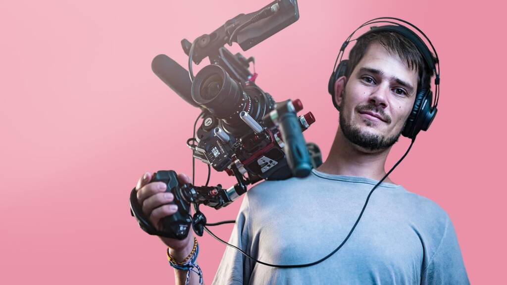 Mann mit Kopfhörern, der eine professionelle Videokamera auf der Schulter trägt und auf einen Betrachter mit rosa Hintergrund blickt.