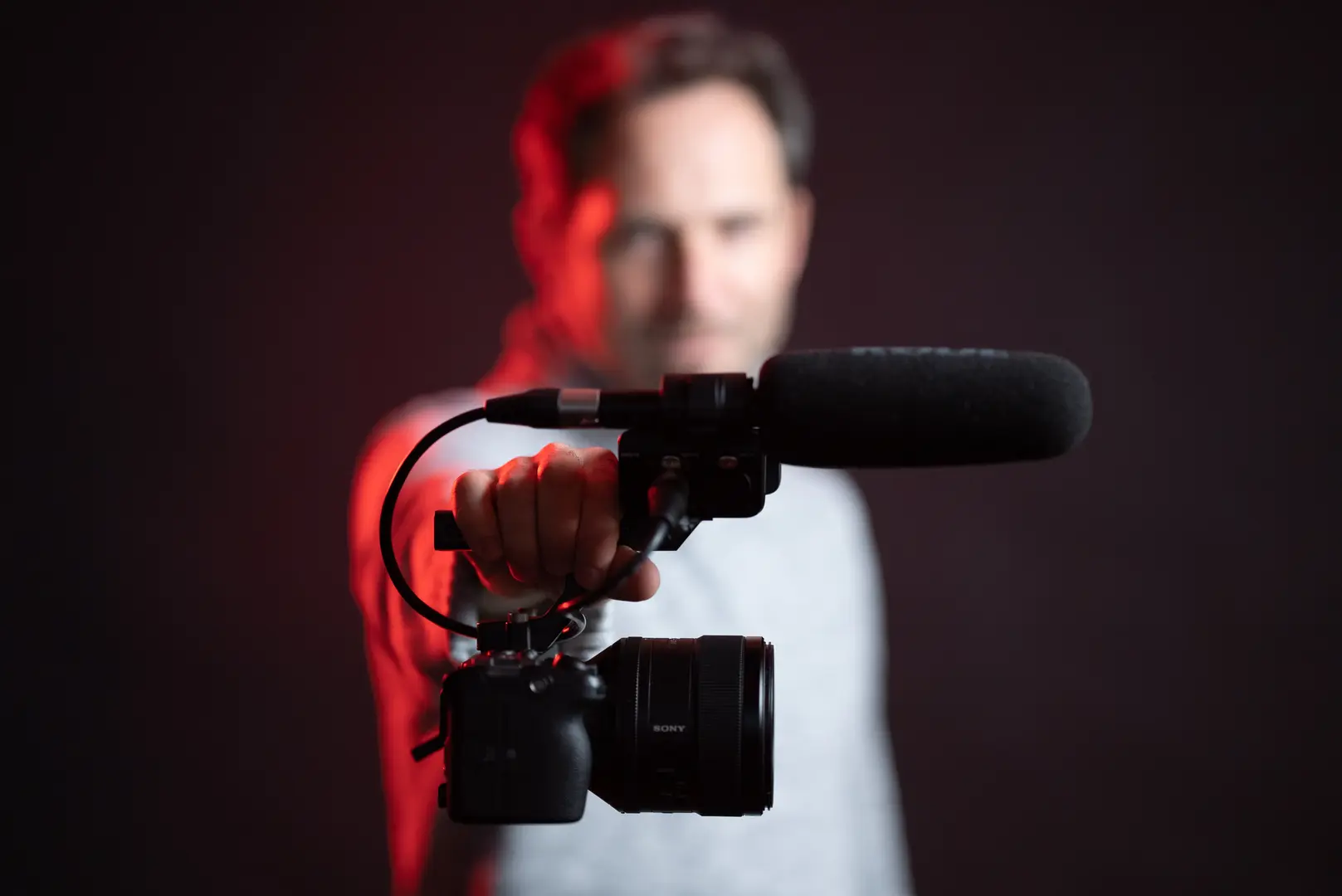 Person hält eine Sony-Videokamera mit angebautem Richtmikrofon in die Kamera, maschinenlesbare Kennzeichnung nach EU AI Act muss direkt in der Datei verankert sein. Rote Beleuchtung von links, dunkler Hintergrund.
