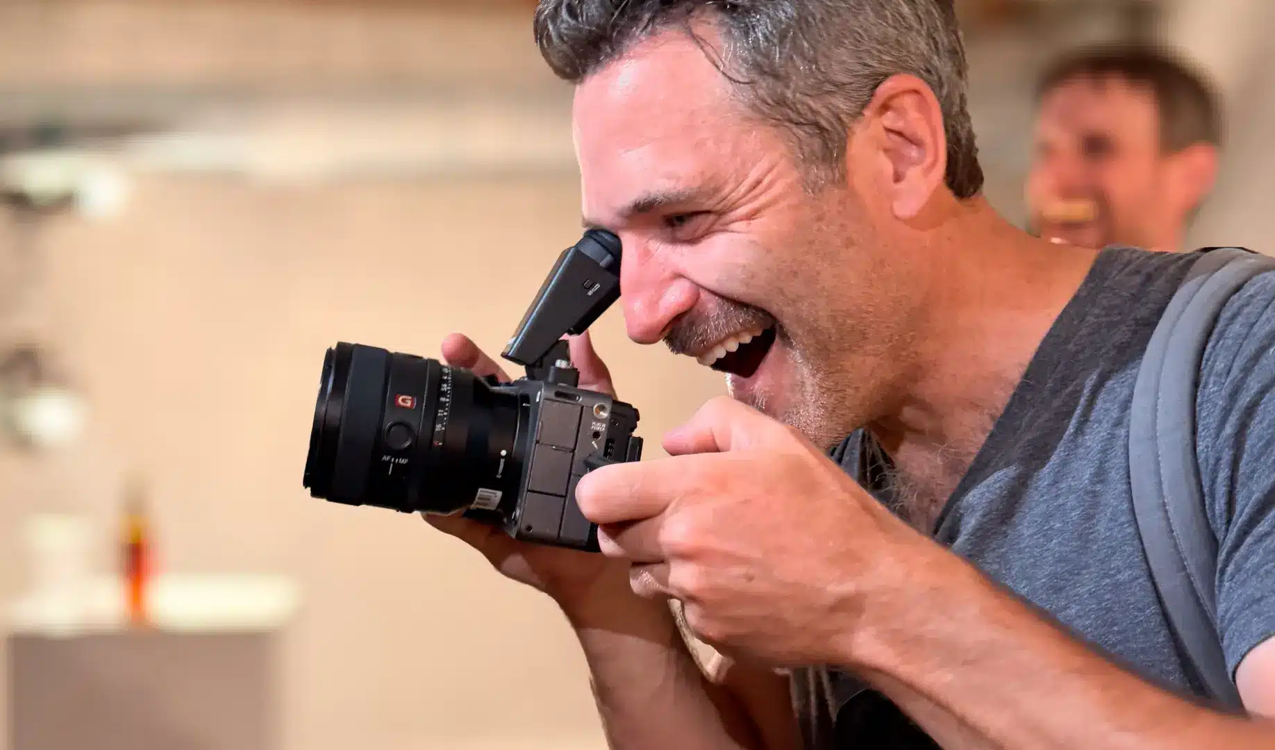 Mann mit Bart hält Sony FX5 Kamera ans Auge, EVF-Monitor ausgeklappt. Er schaut durch den elektronischen Sucher, im unscharfen Hintergrund weitere Personen. Graue Umgebung.