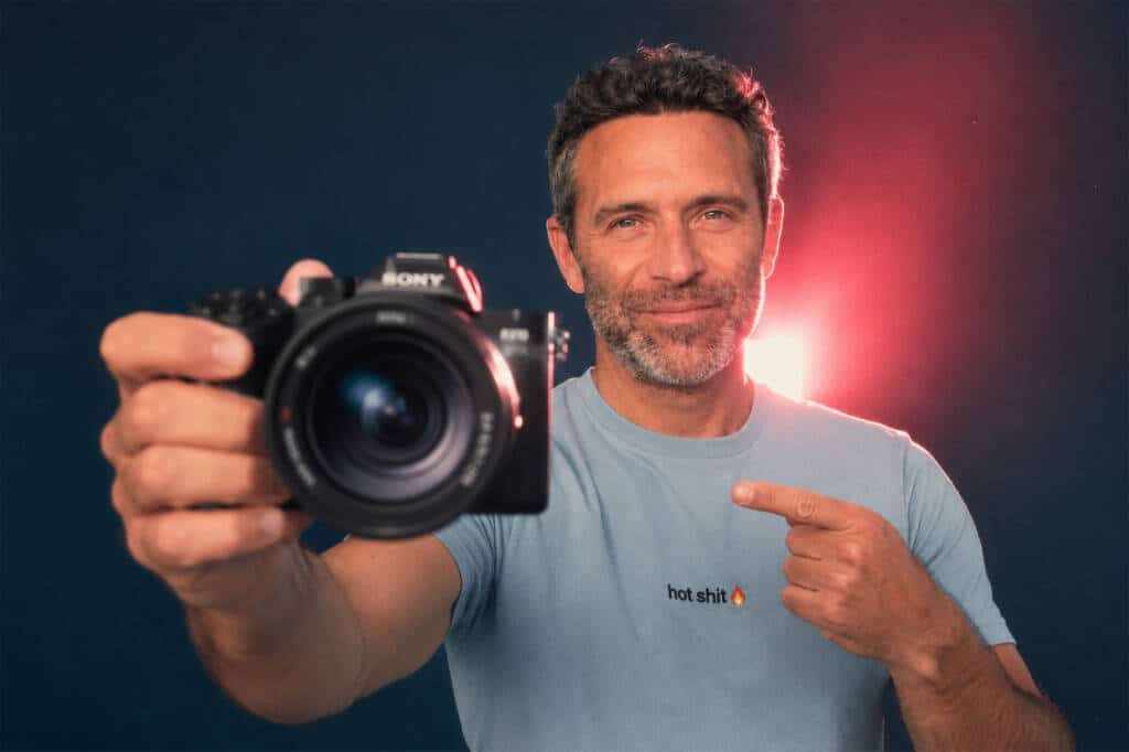 Ein Mann mit kurzen Haaren hält eine Sony RX10 V-Kamera in der Hand und zeigt auf sein hellblaues T-Shirt, auf dem „hot shit“ zusammen mit einem Flammen-Emoji steht, während hinter ihm ein rosafarbenes Licht zu sehen ist.