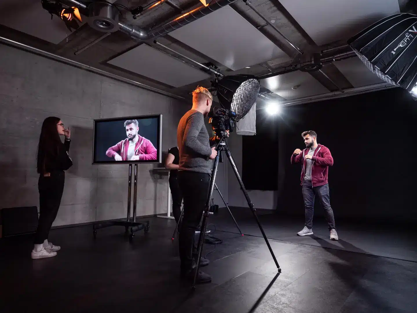 Videostudio-Szenario mit vier Personen: Eine Frau beobachtet von links, ein Mann sitzt vor einer Kamera auf Stativ, eine zweite Person operiert die Kamera, rechts steht ein Mann in roter Jacke vor einem Monitor mit Live-Feed. Decke mit Beleuchtung und Overhead-Struktur sichtbar.