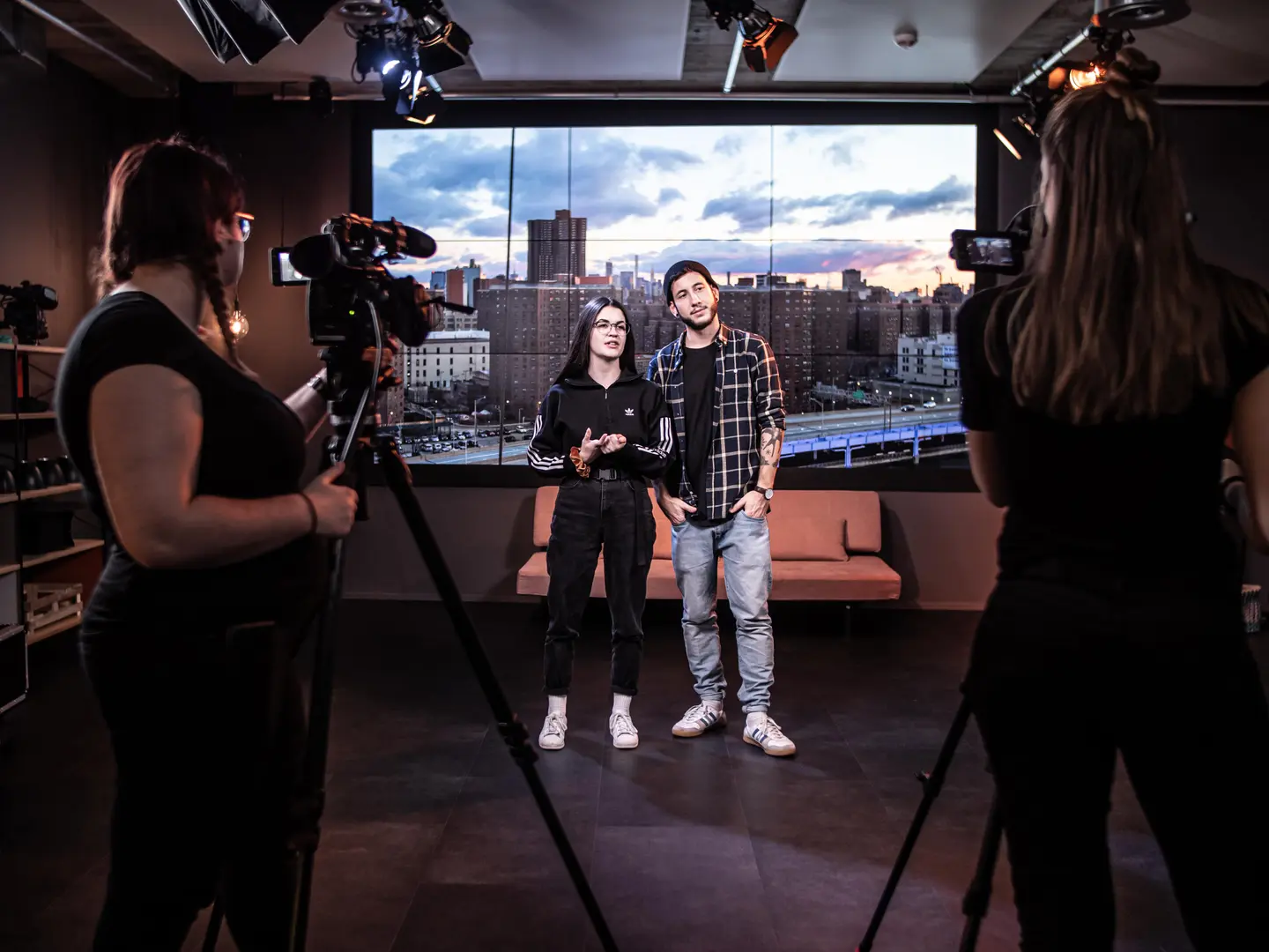Zwei Personen stehen sich in einem TV-Studio gegenüber. Links und rechts je eine Kamerafrau mit Filmkameras auf Stativen. Im Hintergrund großer Monitor mit Stadtpanorama bei Sonnenuntergang. Orange Sofa, professionelle Beleuchtung von oben.