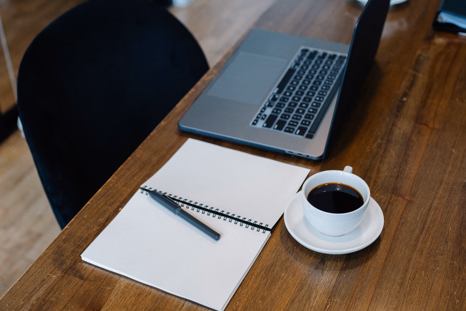 Holztisch mit aufgeklapptem Laptop, spiralgebundenem Notizbuch, Kugelschreiber und weißer Kaffeetasse auf Untertasse. Schwarzer Stuhl im Hintergrund.