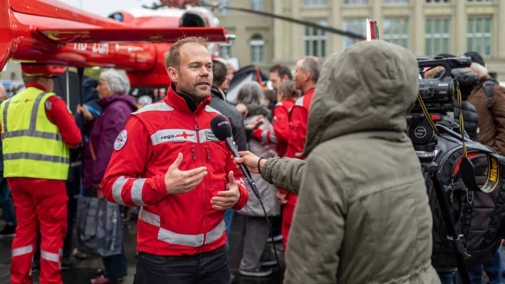 Adrian Schindler in roter Rega-Mediensprecher-Jacke gibt einem Kamerateam ein Interview, im Hintergrund ein Rettungshelikopter und Besucher einer Veranstaltung.
