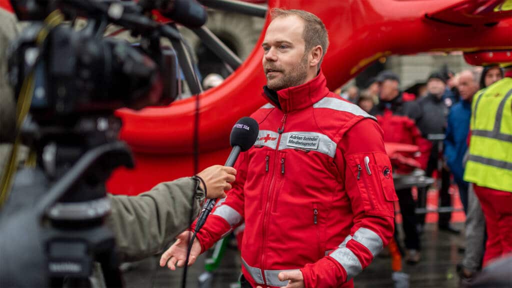 Adrian Schindler in roter Rettungsuniform wird vor einem Hubschrauber vor der Kamera interviewt; er hält eine Checkliste in der Hand, im Hintergrund sind Rettungskräfte zu sehen.