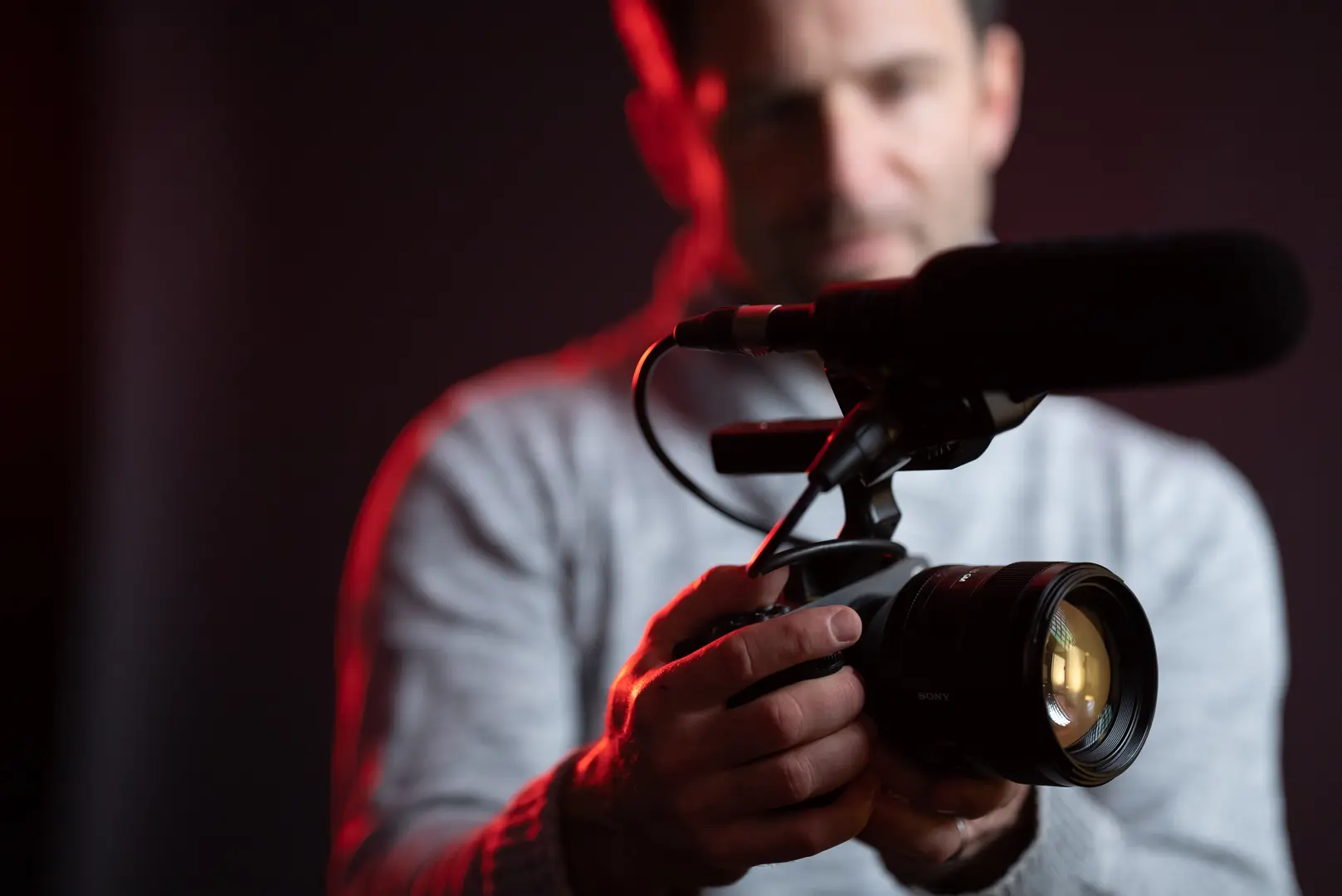 Eine Person hält eine Mirrorless-Kamera mit großem Objektiv und aufgesetztem Richtmikrofon vor dunklem Hintergrund; ein Stativ ist nicht sichtbar.