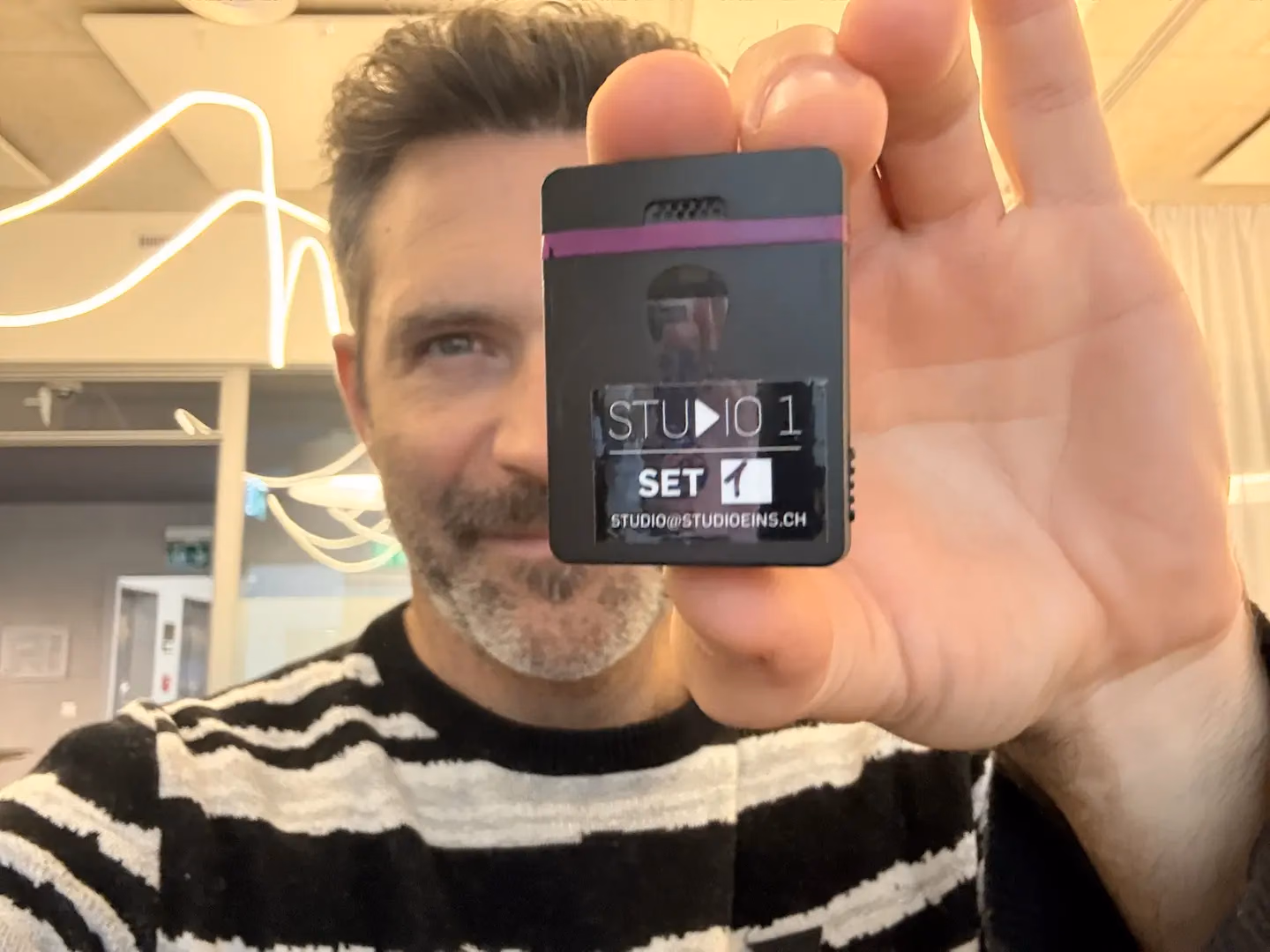 Roman Lehmann hält einen schwarzen Tentacle-Timecode-Generator mit violettem Band und dem Etikett «Studio 1 Set 1» in die Kamera.